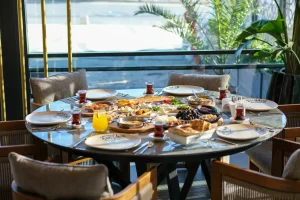 Ekmek, peynir, meyve ve çay gibi çeşitli yemeklerin bulunduğu kahvaltı için hazırlanmış yuvarlak bir yemek masası. Çakı Restorandaki büyük bir pencereden süzülen güneş ışığı, arka planda sandalyeler ve saksı bitkileriyle çevrili masayı aydınlatıyor.